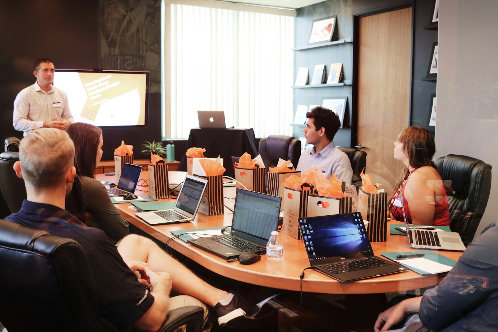A presenter briefing a seated group around a conference table with laptops and gift bags