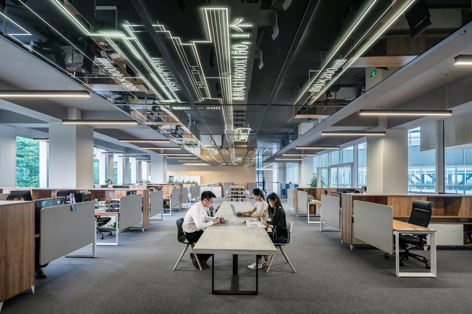 An open-plan office with desk rows along both sides and three colleagues working at a shared central table