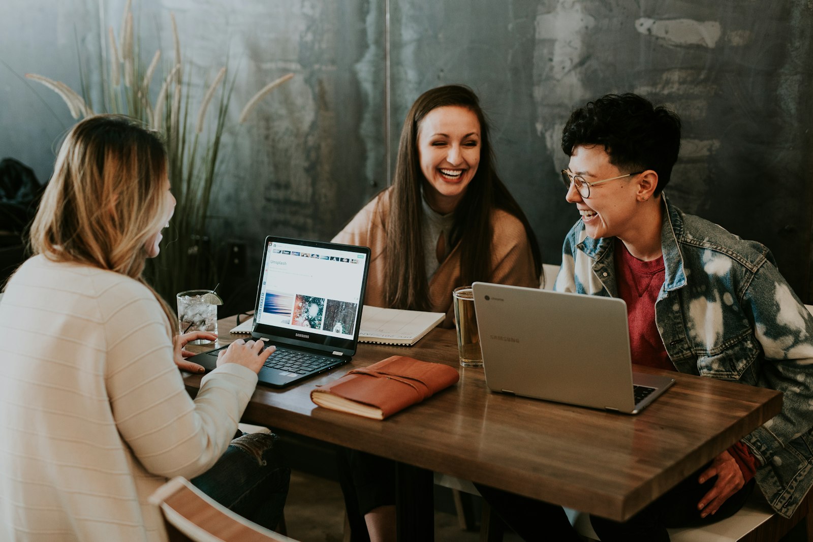 Three colleagues laughing together at a wooden table with laptops and drinks