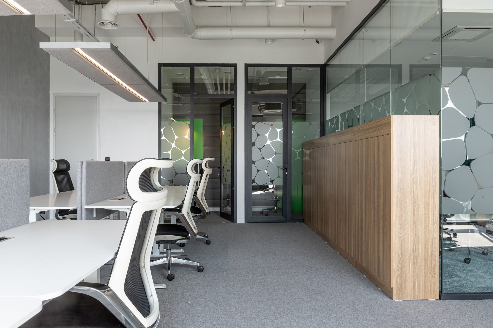 A glass walled corridor with a lounge area visible through the partitions, furnished with a sofa and a floor lamp.