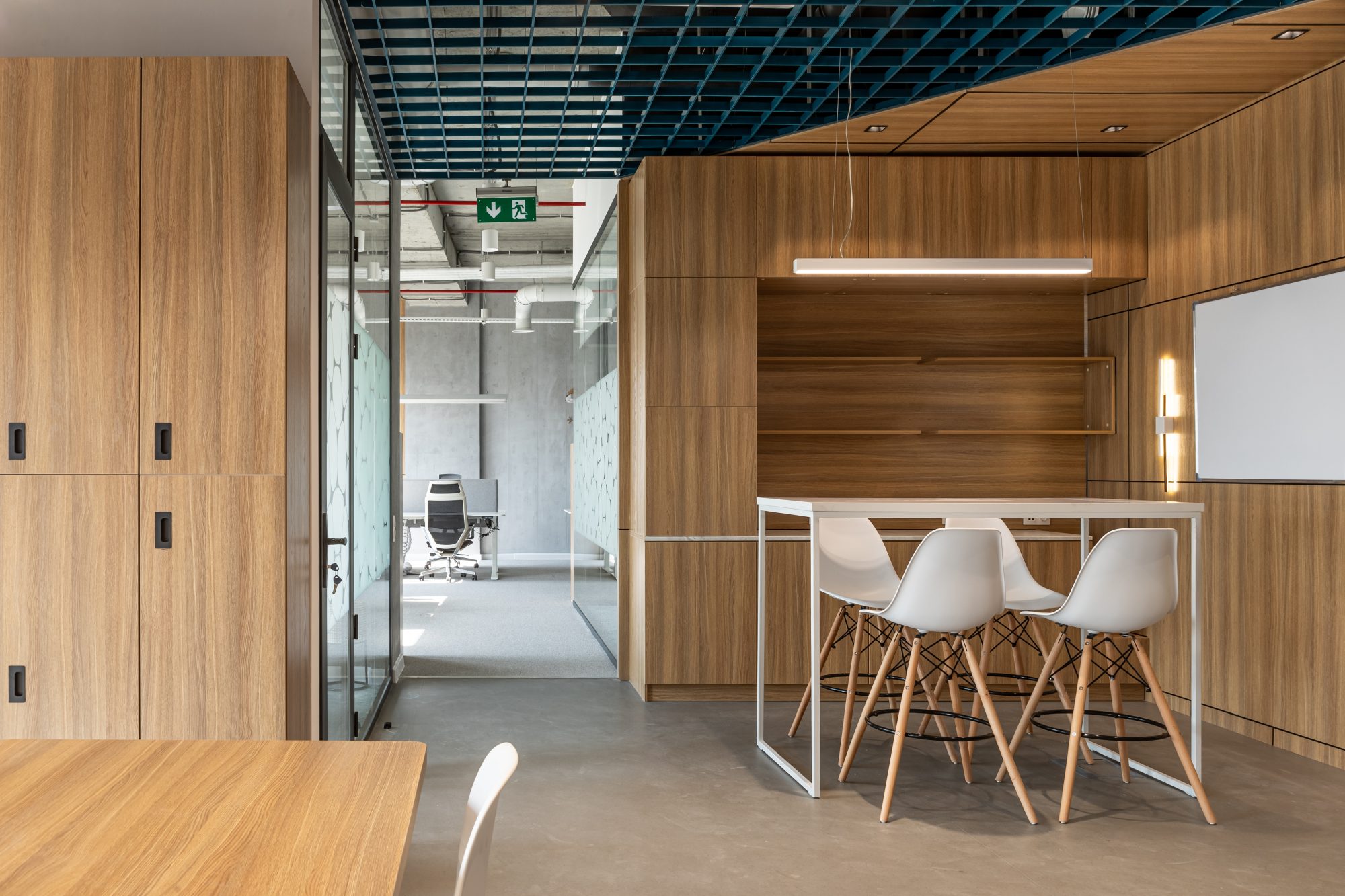 An empty break out area with a bench, low shelving and a kitchenette beside a glass walled corridor.